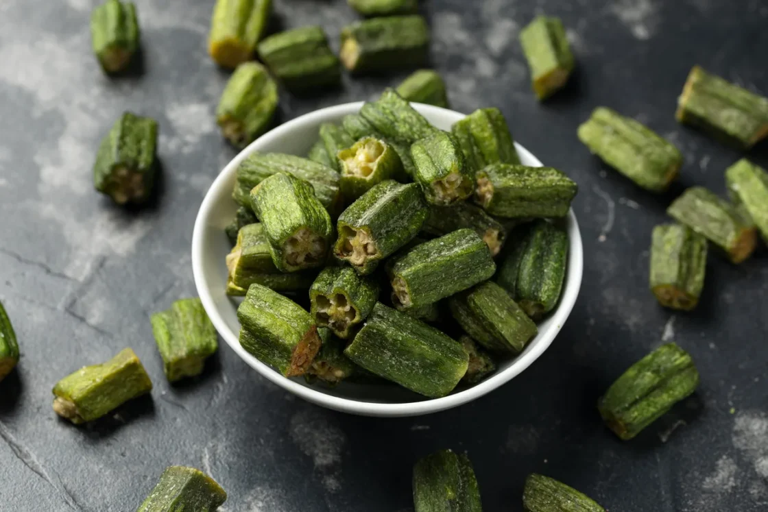 Fresh green okra pods displayed in a white bowl with scattered pieces on dark surface, ideal for commercial food suppliers and wholesalers.