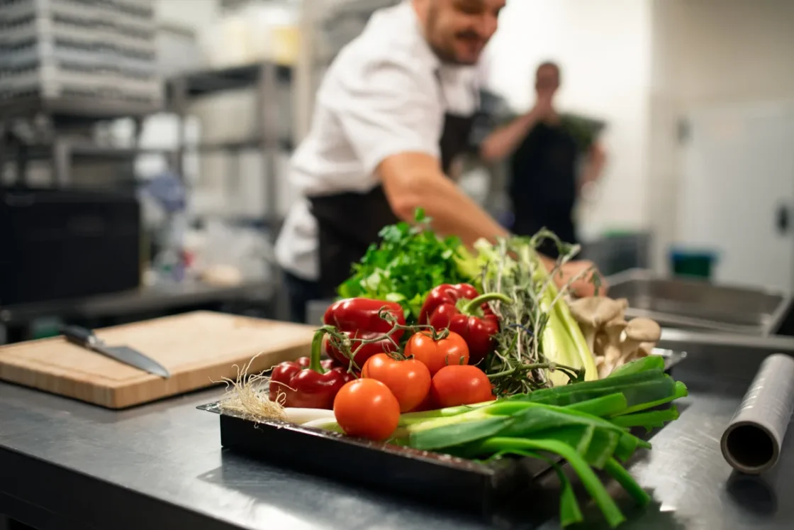 Chef arranges fresh vegetables including tomatoes, peppers, and greens on a prep station in a professional commercial kitchen setting.