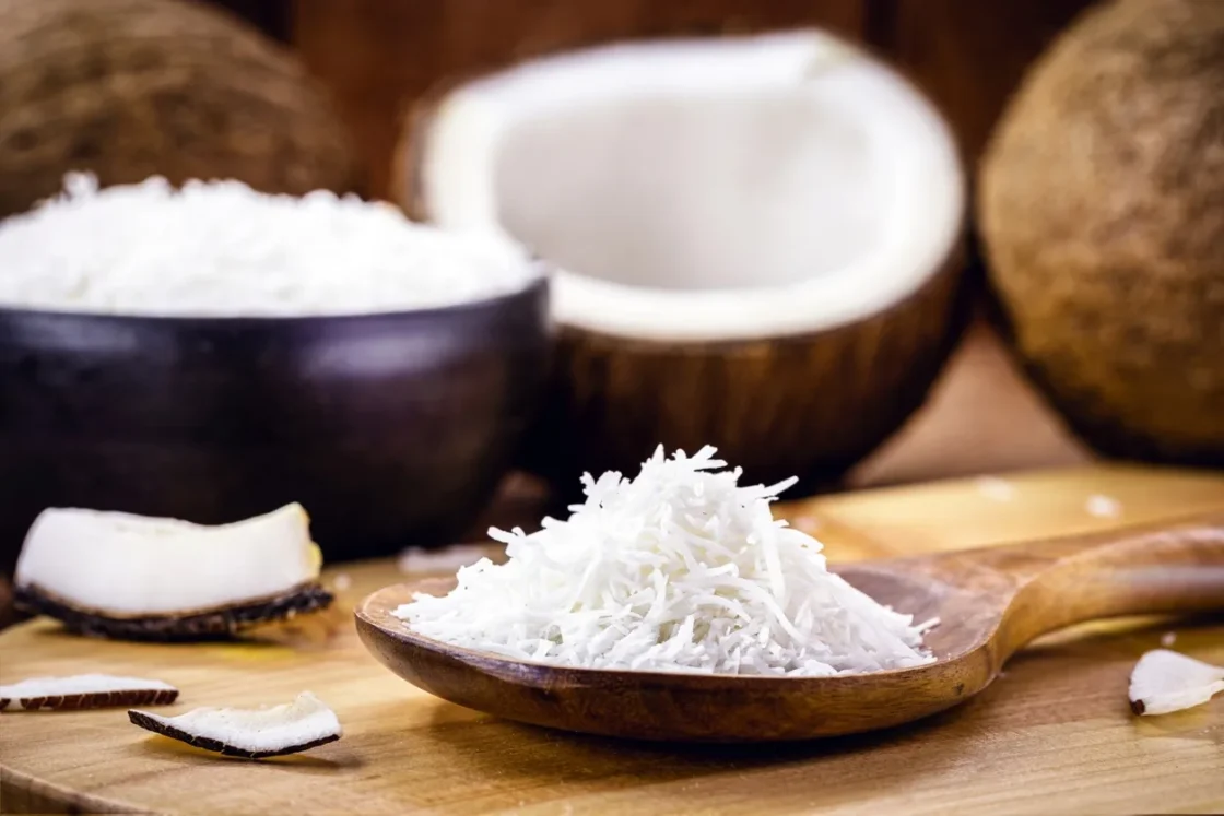 Shredded coconut displayed on wooden spoon with fresh coconut halves, ideal for food manufacturers and distributors.